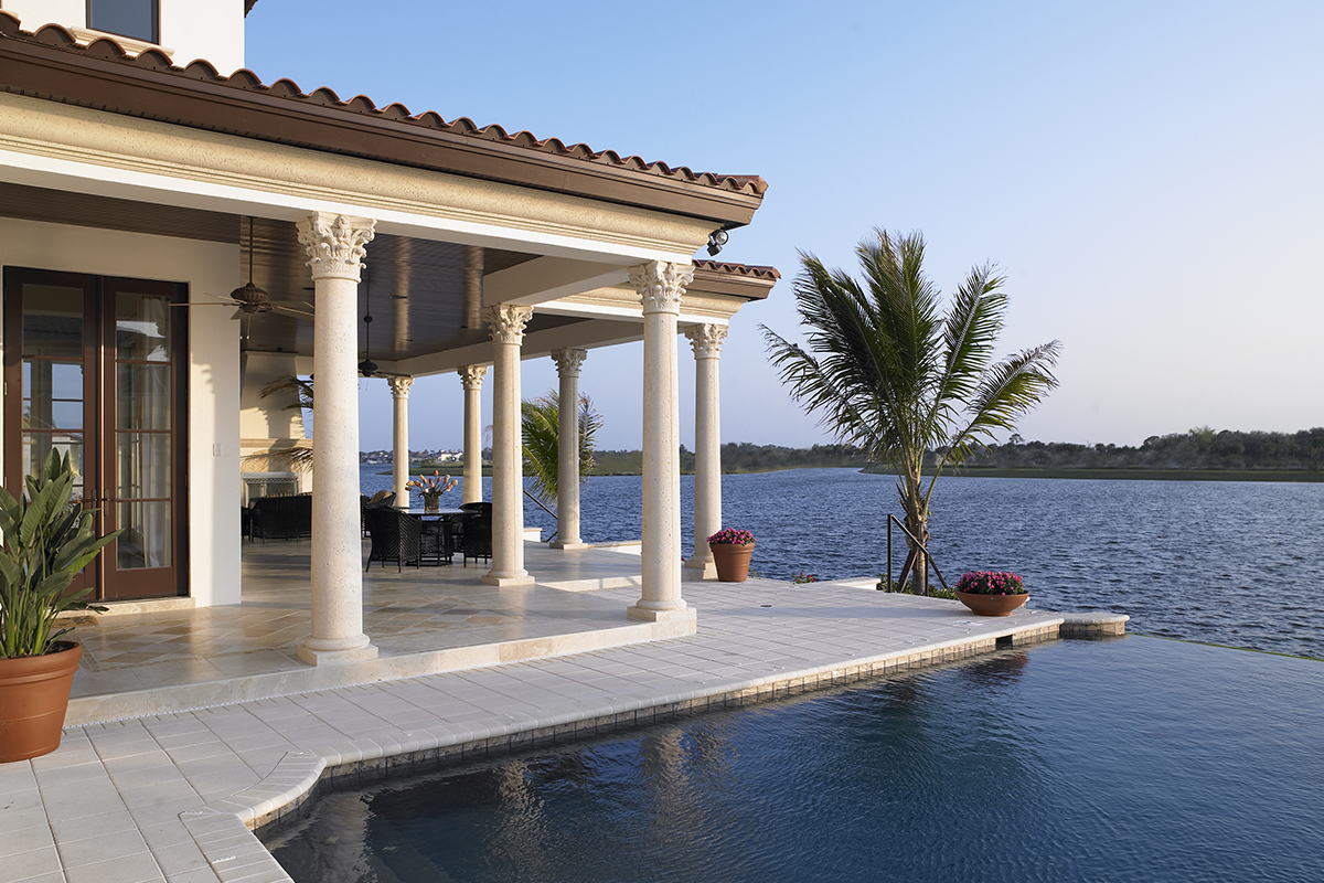 A Florida waterfront home with a columned lanai, tile roof and an infinity pool looking out over open water at dusk.