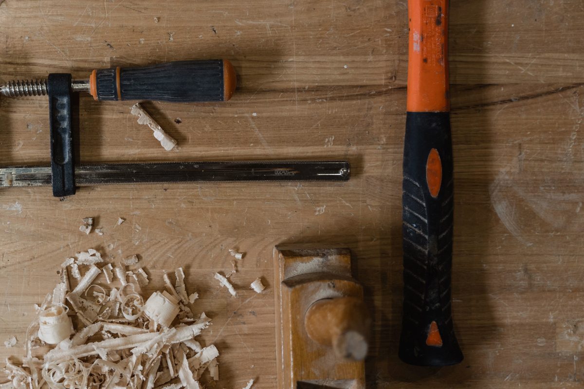 A hammer, clamp, file and wooden hand plane laid out on a worn workbench beside a pile of wood shavings.