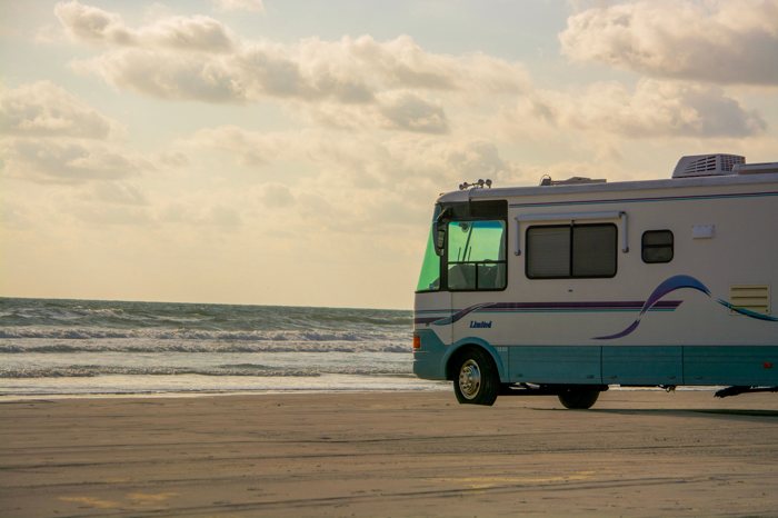 A large American Class A motorhome parked on an empty stretch of beach at golden hour, facing the surf.